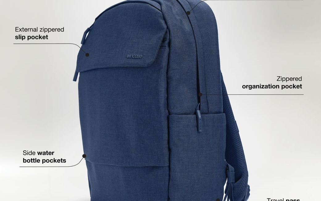 Oblique view of the incase Crosstown Backpack which is medium blue in color with a denim like material. Called out in the photo are an external zippered slip pocket, a padded faux fur laptop compartment for up to 16” devices, a zippered organization pocket, side water bottle pockets, and a travel pass through. One of the two backpacks straps can be seen emerging from the rear.